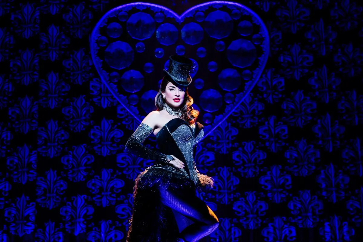 Satine con sombrero de copa y corsé de pedrería ante un corazón de cristal iluminado en azul